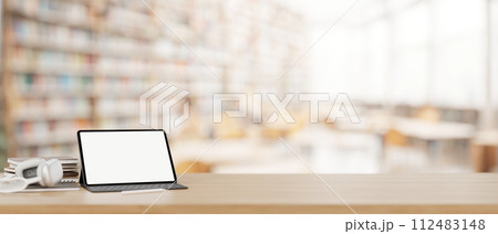 A tablet mockup, headphones, and books on a wooden desk with a blurred background of a library. 112483148