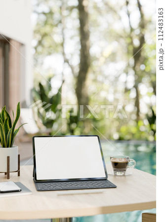 A digital tablet mockup, a wireless keyboard, a coffee cup, and decor on a table by the pool. A digital tablet mockup, a wireless keyboard, a coffee cup, and decor on a table by the pool. 112483163