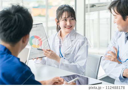 医局のカンファレンス・ミーティングで資料・グラフを用いて説明・提案する白衣姿の女性 112488178