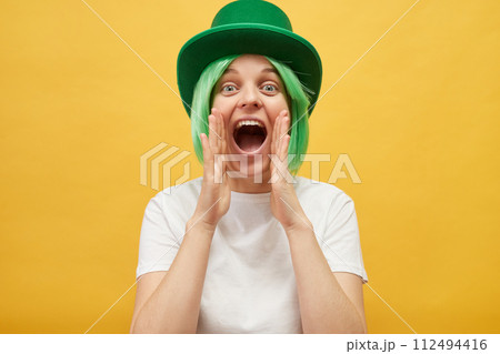 Inviting for St. Patrick's day party. Excited Caucasian woman wearing green leprechaun hat standing isolated over yellow background screaming with hands near mouth making announce 112494416