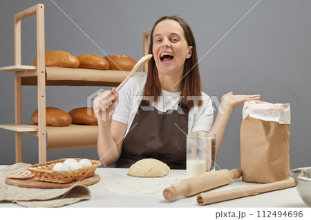 Excited woman wearing brown apron kneading yeast dough and having fun holding wooden spoon as microphone singing while baking bread in bakery Excited woman wearing brown apron kneading yeast dough and having fun holding wooden spoon as microphone singing while baking bread in bakery 112494696