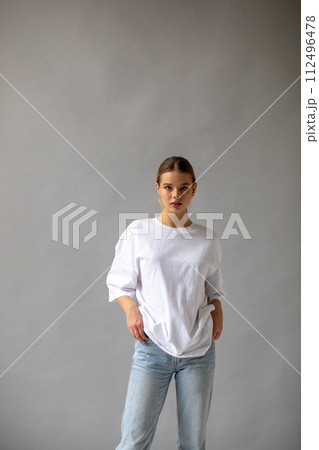 Beautiful blonde girl in a white oversized T-shirt and blue jeans posing on a gray background Beautiful blonde girl in a white oversized T-shirt and blue jeans posing on a gray background 112496478