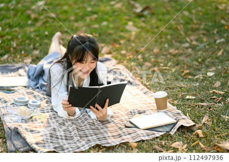 Relaxed young woman lying on picnic blanket and reading boo, enjoying free time at outdoor 112497936