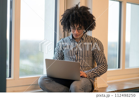 Curly-haired man sitting on a window-sill and working on laptop Curly-haired man sitting on a window-sill and working on laptop 112498250