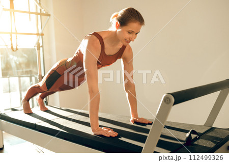 Slim woman wearing sports attire having anaerobic exercise on reformer Slim woman wearing sports attire having anaerobic exercise on reformer 112499326