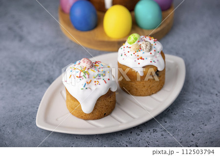 Two small Easter kulichs with candied fruits in white glaze with colorful sprinkles in the cut. Painted chicken and quail eggs. Traditional Easter baking. Easter holiday. Close-up, selective focus. Two small Easter kulichs with candied fruits in white glaze with colorful sprinkles in the cut. Painted chicken and quail eggs. Traditional Easter baking. Easter holiday. Close-up, selective focus. 112503794