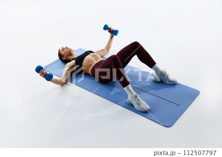 Young woman with slim, fit body in sportswear training on fitness mat, lifting dumbbells, isolated over white studio background 112507797