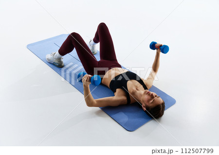 Young woman with slim, fit body in sportswear training on fitness mat, lifting dumbbells, isolated over white studio background 112507799