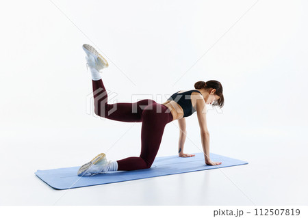 Young athletic woman in leggings and top training, standing on knees and plans and lifting leg isolated over white studio background 112507819