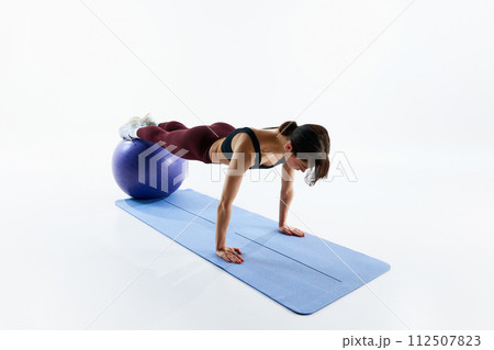 Sportive young woman with fit body training, standing in plank position on fitness ball isolated over white studio background 112507823