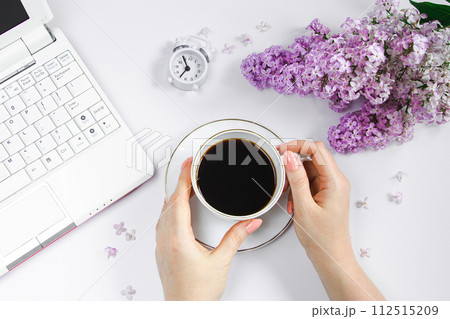 Female hands holding cup of espresso coffee. Alarm clock and lilac flowers on light background. 112515209