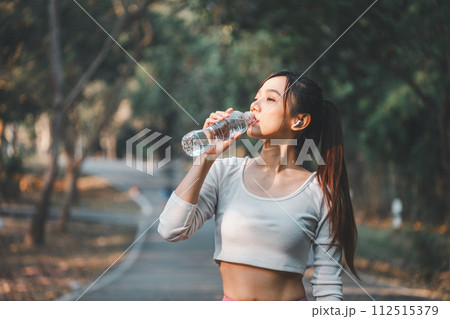 A health-conscious woman takes a refreshing break to drink water during her outdoor exercise routine, highlighting the importance of hydration in fitness. 112515379