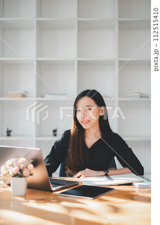 A young, confident professional woman sits at her desk in a minimalist office, working on her laptop with a tablet and notebook at hand, surrounded by the clean lines and simplicity of a modern 112515410