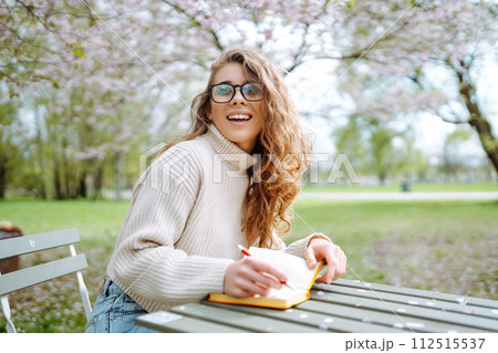 Young woman sitting at a table in blooming spring park with a notepad and laptop. Lifestyle concept. Young woman sitting at a table in blooming spring park with a notepad and laptop. Lifestyle concept. 112515537