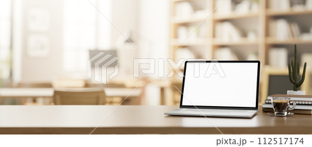 A wooden table features a laptop mockup in a cosy neutral coffee shop or library co-working space. 112517174