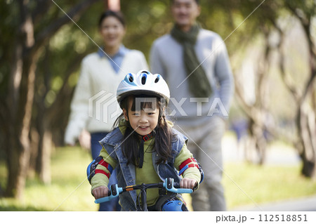 little asian girl riding bike in city park with parents in background 112518851