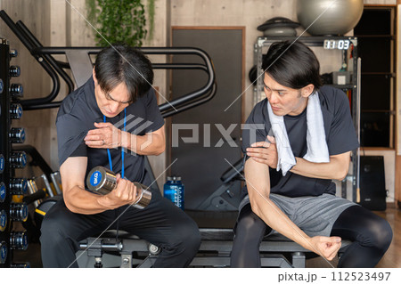 パーソナルジムで会員にトレーニングの見本を見せるトレーナーの男性(腕トレ) パーソナルジムで会員にトレーニングの見本を見せるトレーナーの男性(腕トレ) 112523497
