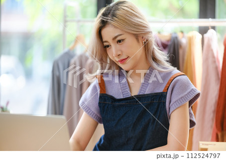 Asian Fashion designer woman using laptop in modern studio the clothes hanging on the racks morning light. 112524797
