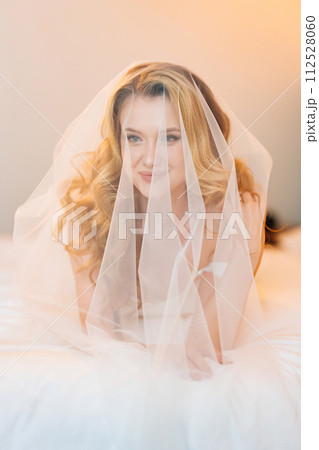 Morning of the bride. Delicate and beautiful blonde bride in bed under veil. 112528060