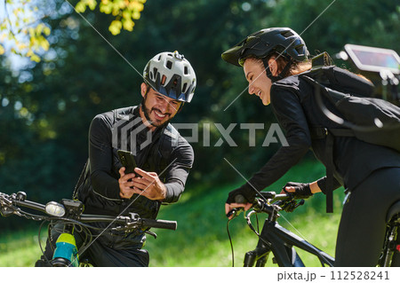 A sweet couple, equipped with bicycles and engrossed in coordinating their journey, checks their GPS mobile and watches while planning scenic routes in the park, seamlessly blending technology and 112528241