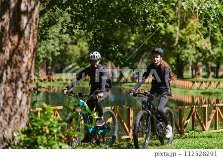 A blissful couple, adorned in professional cycling gear, enjoys a romantic bicycle ride through a park, surrounded by modern natural attractions, radiating love and happiness 112528291