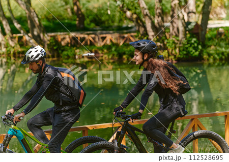 A blissful couple, adorned in professional cycling gear, enjoys a romantic bicycle ride through a park, surrounded by modern natural attractions, radiating love and happiness A blissful couple, adorned in professional cycling gear, enjoys a romantic bicycle ride through a park, surrounded by modern natural attractions, radiating love and happiness 112528695