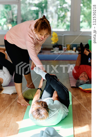 A skilled trainer oversees a group of senior women practicing various yoga exercises, including neck, back, and leg stretches, in a sunlit space, promoting wellness and harmony A skilled trainer oversees a group of senior women practicing various yoga exercises, including neck, back, and leg stretches, in a sunlit space, promoting wellness and harmony 112530218