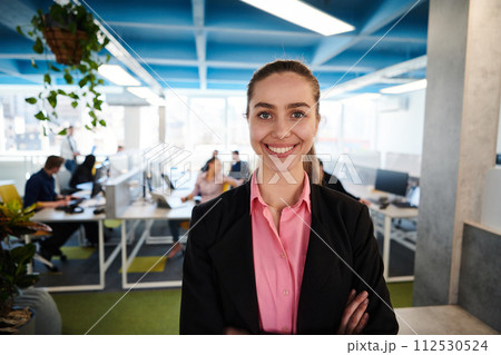 Portrait of young smiling business woman in creative open space coworking startup office. Successful businesswoman standing in office with copyspace. Coworkers working in background 112530524