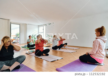 A group of senior women engage in various yoga exercises, including neck, back, and leg stretches, under the guidance of a trainer in a sunlit space, promoting well-being and harmony A group of senior women engage in various yoga exercises, including neck, back, and leg stretches, under the guidance of a trainer in a sunlit space, promoting well-being and harmony 112532192