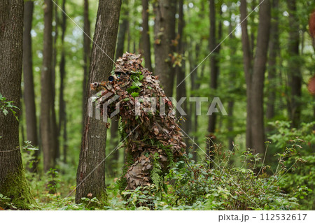 A highly skilled elite sniper, camouflaged in the dense forest, stealthily maneuvers through dangerous woodland terrain on a covert and precise mission 112532617