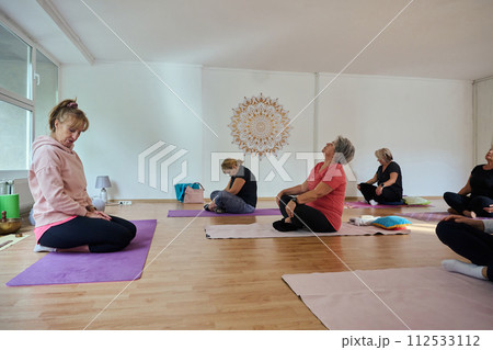 A group of senior women engage in various yoga exercises, including neck, back, and leg stretches, under the guidance of a trainer in a sunlit space, promoting well-being and harmony A group of senior women engage in various yoga exercises, including neck, back, and leg stretches, under the guidance of a trainer in a sunlit space, promoting well-being and harmony 112533112