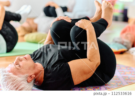 A group of senior women engage in various yoga exercises, including neck, back, and leg stretches, under the guidance of a trainer in a sunlit space, promoting well-being and harmony 112533616