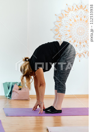 In a sunlit space, a senior woman gracefully practices rejuvenating yoga, focusing on neck, back, and leg stretches, embodying serenity and well-being 112533938