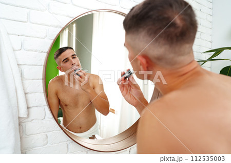 Handsome young man shaving face with electric razor on white background 112535003