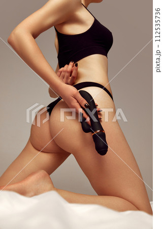 Cropped image of female body, buttocks in black underwear. Woman on bed holding vibrator toy against beige studio background Cropped image of female body, buttocks in black underwear. Woman on bed holding vibrator toy against beige studio background 112535736