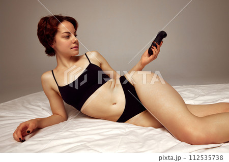 Young woman in black underwear lying on bed, holing vibrator against beige studio background Young woman in black underwear lying on bed, holing vibrator against beige studio background 112535738