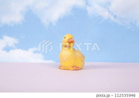 a plaster figure of a yellow chick on a pink ground and in front of a blue sky background with clouds a plaster figure of a yellow chick on a pink ground and in front of a blue sky background with clouds 112535946