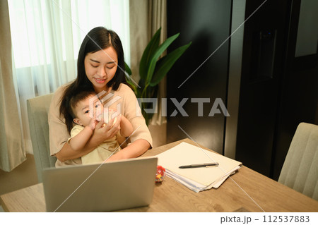 Caring mother feeding milk from bottle to her baby while working remotely at home Caring mother feeding milk from bottle to her baby while working remotely at home 112537883