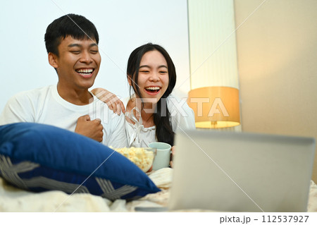 Happy young couple watching movie on laptop and eating popcorn in their bedroom Happy young couple watching movie on laptop and eating popcorn in their bedroom 112537927