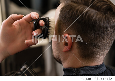 A barber sweeps hair with brush while trimming customers beards with an electric trimmer in the salon. A barber sweeps hair with brush while trimming customers beards with an electric trimmer in the salon. 112538412