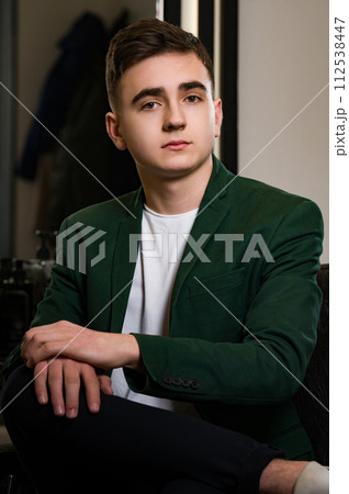 Portrait of a young barber in a jacket and white t-shirt. The client is sitting in a barbershop chair after a haircut, satisfied with the result. 112538447