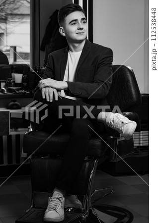 Portrait of a young barber in a jacket and white t-shirt. The client is sitting in a barbershop chair after a haircut, satisfied with the result. Portrait of a young barber in a jacket and white t-shirt. The client is sitting in a barbershop chair after a haircut, satisfied with the result. 112538448