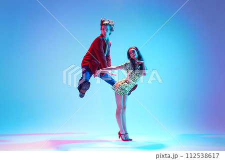 Dynamic image of young artistic couple dancing retro dance, performing against blue background in neon light 112538617
