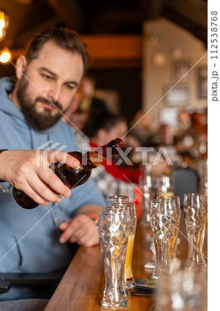A young handsome man with a beard pouring beer from bottle into glasses 112538768