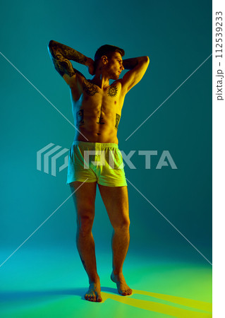 Young man with muscular, athletic body posing raising arms holding behind head in neon light against gradient studio background. 112539233