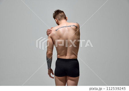 Rear view of young man with tattoos on body holds hand on shoulder shows pain against grey studio background. Copy space. 112539245