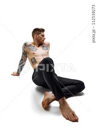 Tattooed, handsome man wearing black pants sitting, posing against white studio background. Copy space. 112539273