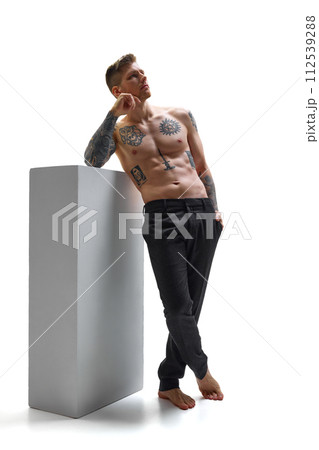 Barefoot, tattooed young man leaning on gray block posing deep in thought against white studio background. Copy space. Barefoot, tattooed young man leaning on gray block posing deep in thought against white studio background. Copy space. 112539288