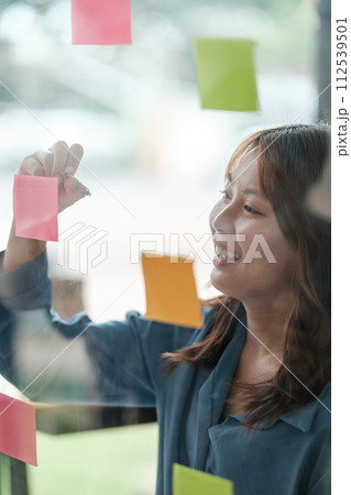 Beautiful female employee write down on colorful sticky notes manage list, concentrated biracial woman work on startup brainstorm collaborate plan on stickers on glass wall. Beautiful female employee write down on colorful sticky notes manage list, concentrated biracial woman work on startup brainstorm collaborate plan on stickers on glass wall. 112539501