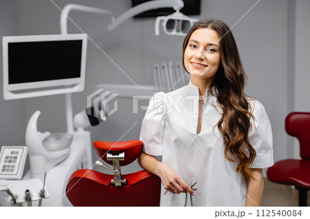 A young pretty female dentist is standing near the dental chair in the office, holding tools for work and looking into the camera A young pretty female dentist is standing near the dental chair in the office, holding tools for work and looking into the camera 112540084